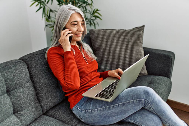 Седовласая женщина средних лет разговаривает на смартфоне с помощью ноутбука дома - Фото, изображение