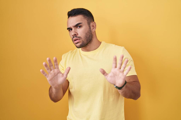 Молодий іспаномовний чоловік стоїть над жовтим тлом, відсуває долоні, показуючи відмову і заперечуючи з страхом і огидною експресією. зупинити і заборонити
.  - Фото, зображення