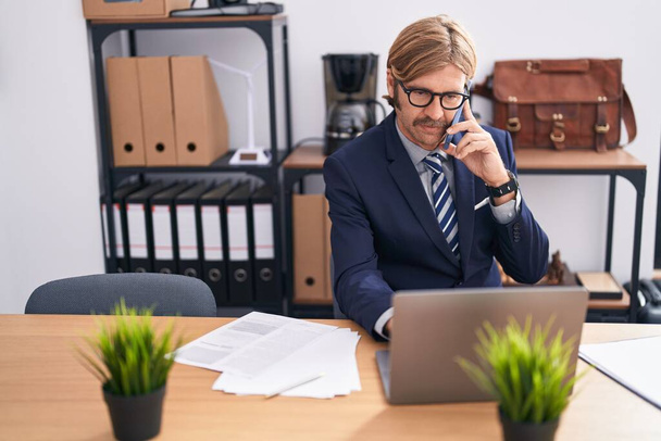 Fiatal szőke férfi üzleti dolgozó laptoppal beszél okostelefonon az irodában - Fotó, kép