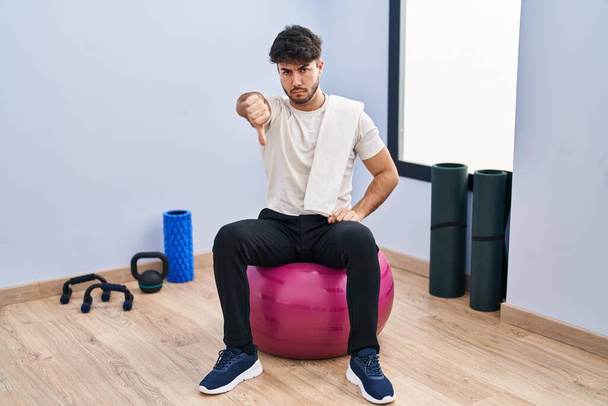 Hispanic man met baard zittend op pilates ballen in yoga kamer kijken ongelukkig en boos tonen afwijzing en negatief met duimen naar beneden gebaar. slechte uitdrukking.  - Foto, afbeelding