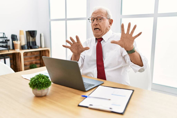 Senior man working at the office using computer laptop afraid and terrified with fear expression stop gesture with hands, shouting in shock. panic concept.  - Photo, image
