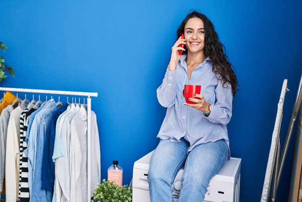 Junge hispanische Frau trinkt Kaffee und telefoniert mit dem Smartphone und wartet in der Waschküche auf die Waschmaschine - Foto, Bild