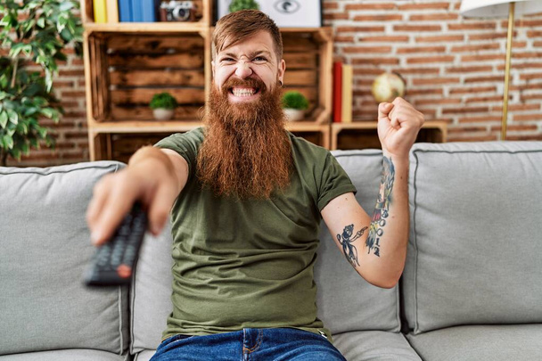 Homme rousse à longue barbe tenant télécommande de télévision criant fier, célébrant la victoire et le succès très excité avec le bras levé  - Photo, image