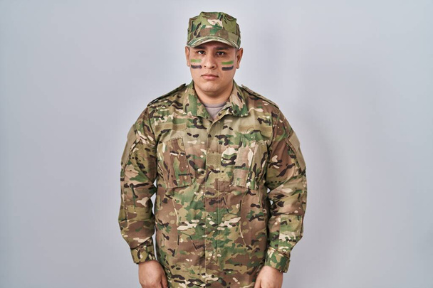 顔に真剣な表情でリラックスしたカモフラージュ軍の制服を着たヒスパニック系の若者。カメラを見ているだけで.  - 写真・画像