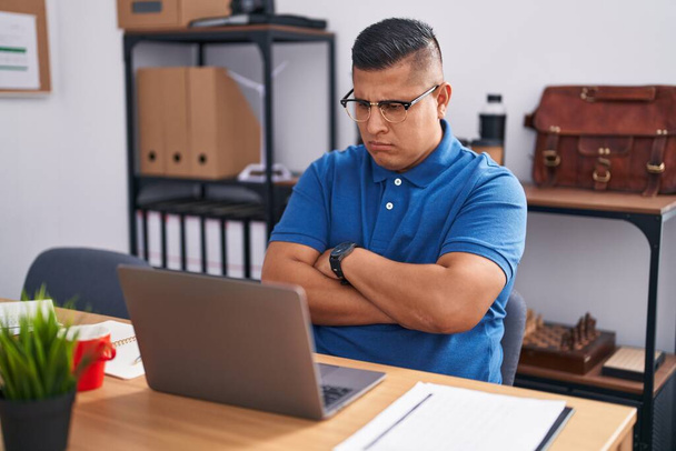 Jovem hispânico trabalhando no escritório com laptop cético e nervoso, desaprovando a expressão no rosto com os braços cruzados. pessoa negativa.  - Foto, Imagem