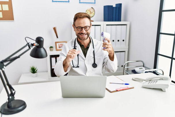 Médico de mediana edad hombre sosteniendo termómetro en la clínica señalando el dedo a uno mismo sonriendo feliz y orgulloso  - Foto, imagen