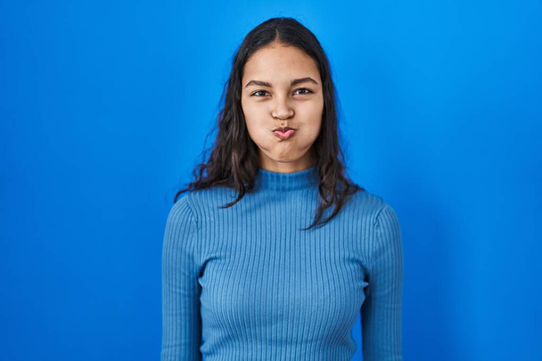 Jeune femme brésilienne debout sur fond bleu isolé joues gonflées avec un visage drôle. bouche gonflée d'air, expression folle.  - Photo, image