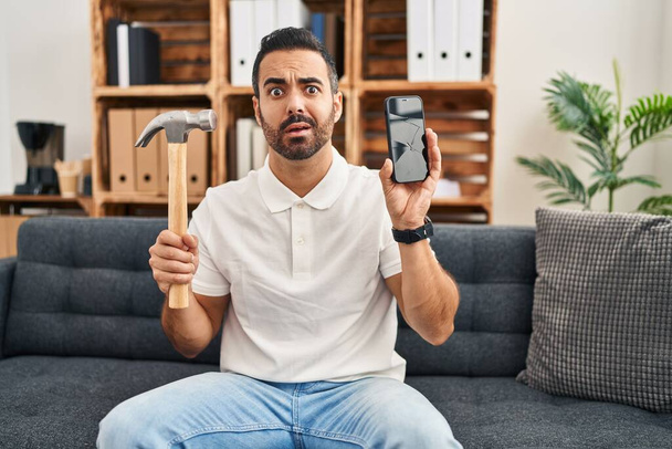 Молодой латиноамериканец с бородой, держа в руках молоток и сломанный смартфон, показывает потрескавшийся экран в шоковом лице, выглядящий скептически и саркастично, удивленный открытым ртом  - Фото, изображение