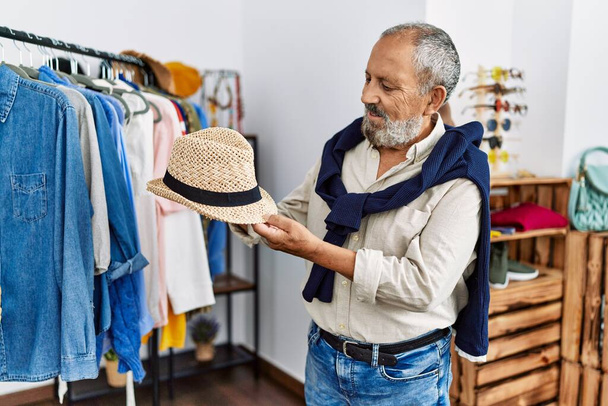 Hombre de pelo gris mayor sonriendo confiado sosteniendo el sombrero en la tienda de ropa - Foto, Imagen