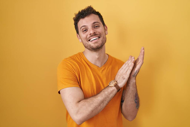 Fiatal spanyol férfi áll sárga háttér tapsol és tapsol boldog és örömteli, mosolygó büszke kezét együtt  - Fotó, kép
