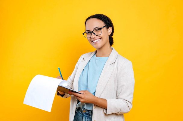 Beautiful young brunette mixed race woman, wearing stylish casual clothes, hr manager, real estate agent, holds folder with papers document, looks at camera and smile, isolated orange background - Fotoğraf, Görsel