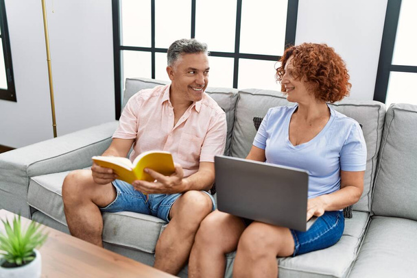 Orta yaşlı erkek ve kadın çift dizüstü bilgisayar kullanıyor ve evde kanepede oturmuş kitap okuyor. - Fotoğraf, Görsel