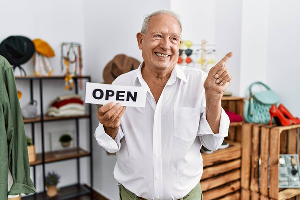 Vanhempi mies pitäen banneria avoimella tekstillä vähittäiskaupassa hymyillen onnellisena osoittaen kädellä ja sormella sivulle  - Valokuva, kuva