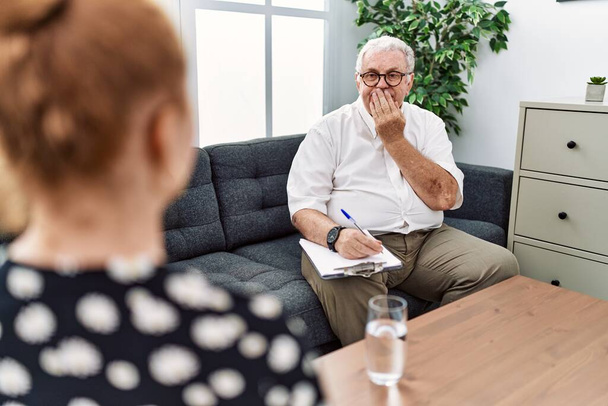 Senior-Psychologe Mann in Beratungsstelle lacht und verlegen kichert Mund mit Händen, Klatsch und Skandalkonzept bedeckt  - Foto, Bild