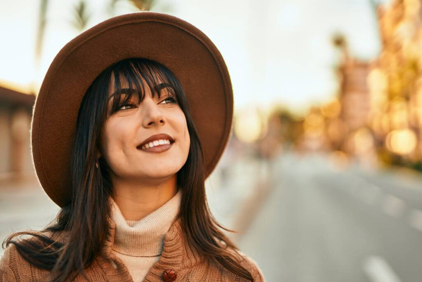 Brunette woman wearing winter hat smiling outdoors at the city on sunset - Photo, image
