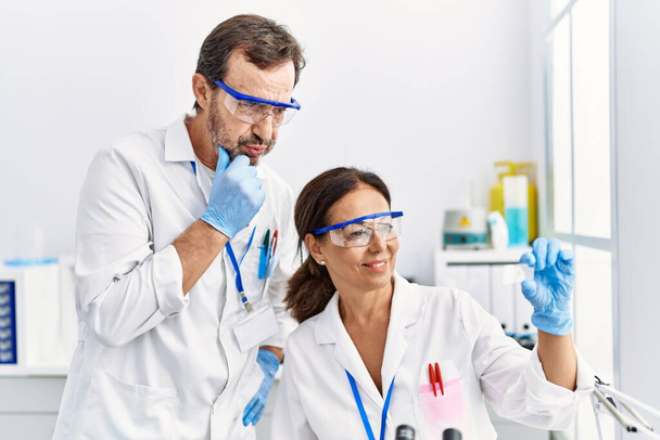Mężczyzna i kobieta w średnim wieku partnerzy w mundurze naukowca analizujący próbki w laboratorium - Zdjęcie, obraz