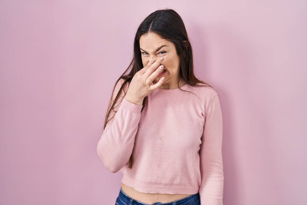 Νεαρή μελαχρινή γυναίκα στέκεται πάνω από ροζ φόντο μυρίζοντας κάτι βρωμερό και αηδιαστικό, ανυπόφορη μυρωδιά, κρατώντας την αναπνοή με τα δάχτυλα στη μύτη. άσχημη μυρωδιά  - Φωτογραφία, εικόνα