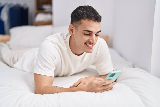Junger hispanischer Mann mit Smartphone im Schlafzimmer liegend - Foto, Bild