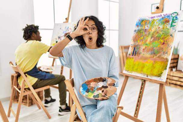 Jonge Latijns-Amerikaanse vrouw in de kunststudio doet ok gebaar geschokt met verrast gezicht, oog kijken door vingers. ongelovige uitdrukking.  - Foto, afbeelding