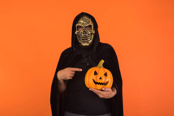 Person mit Totenkopfmaske und schwarzem Kapuzenumhang, die mit dem Finger auf einen Kürbis zeigt, der auf orangefarbenem Hintergrund Halloween feiert. Festkonzept, Allerseelen und Allerheiligen. - Foto, Bild