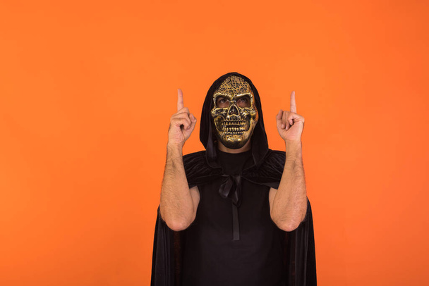 Πρόσωπο με μάσκα κρανίου και μαύρη κάπα με κουκούλα, κοιτάζει και δείχνει τα δάχτυλα προς τα πάνω, γιορτάζει το Halloween, σε πορτοκαλί φόντο. Εορταστική ιδέα, Ημέρα των Ψυχών και Ημέρα των Αγίων Πάντων. - Φωτογραφία, εικόνα