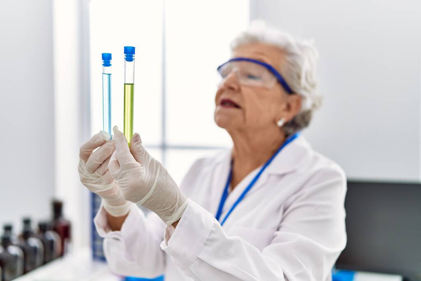 Ηλικιωμένη γκριζομάλλα γυναίκα που φορά στολή επιστήμονα κρατώντας δοκιμαστικούς σωλήνες στο εργαστήριο - Φωτογραφία, εικόνα