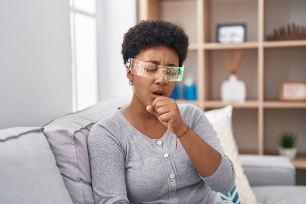 Νεαρή Αφροαμερικανή γυναίκα που φοράει γυαλιά εικονικής πραγματικότητας κάθεται στον καναπέ νιώθοντας αδιαθεσία και βήχει ως σύμπτωμα για το κρύο ή τη βρογχίτιδα. έννοια της υγειονομικής περίθαλψης.  - Φωτογραφία, εικόνα