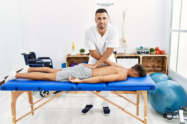 Dos hombres hispanos fisioterapeuta y paciente que tienen sesión de rehabilitación masajeando en la clínica - Foto, imagen
