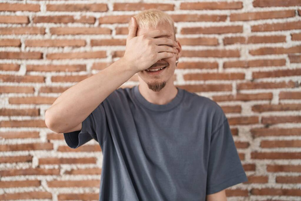 Young caucasian man standing over bricks wall smiling and laughing with hand on face covering eyes for surprise. blind concept.  - Photo, Image