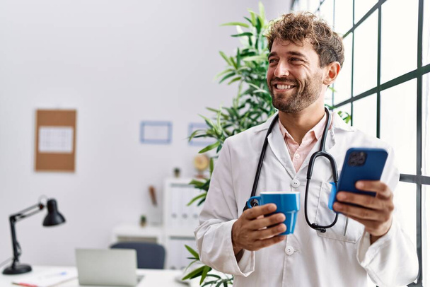 Mladý hispánec muž nosí lékařskou uniformu pomocí smartphonu a pití kávy na klinice - Fotografie, Obrázek