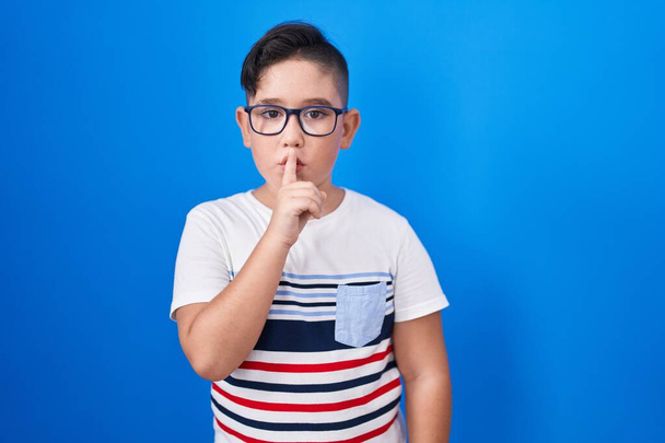 Junge hispanische Kinder, die vor blauem Hintergrund stehen und mit dem Finger auf den Lippen um Ruhe bitten. Schweigen und geheimes Konzept.  - Foto, Bild