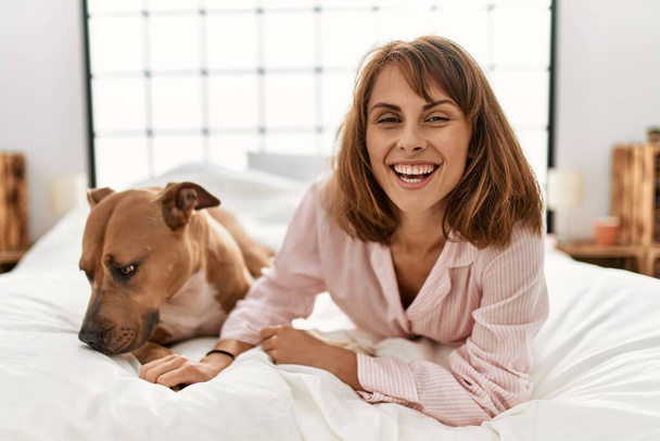 Νεαρή καυκάσια γυναίκα χαμογελά με αυτοπεποίθηση ξαπλωμένη στο κρεβάτι με το σκύλο στο υπνοδωμάτιο - Φωτογραφία, εικόνα