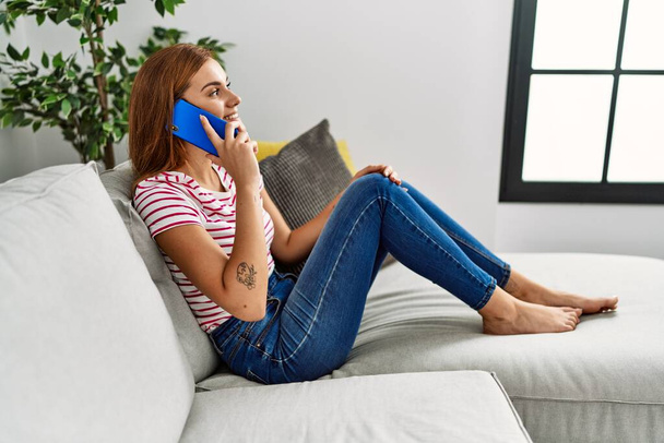 Młoda kobieta rozmawia na smartfonie siedząc na kanapie w domu - Zdjęcie, obraz