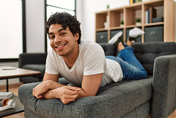 Junger hispanischer Mann lächelt glücklich zu Hause auf dem Sofa liegend. - Foto, Bild