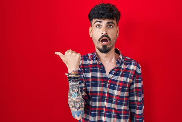 Νεαρός Ισπανόφωνος άνδρας με γενειάδα στέκεται πάνω από το κόκκινο φόντο έκπληκτος δείχνοντας με το χέρι δάχτυλο στο πλάι, ανοιχτό στόμα κατάπληκτος έκφραση.  - Φωτογραφία, εικόνα