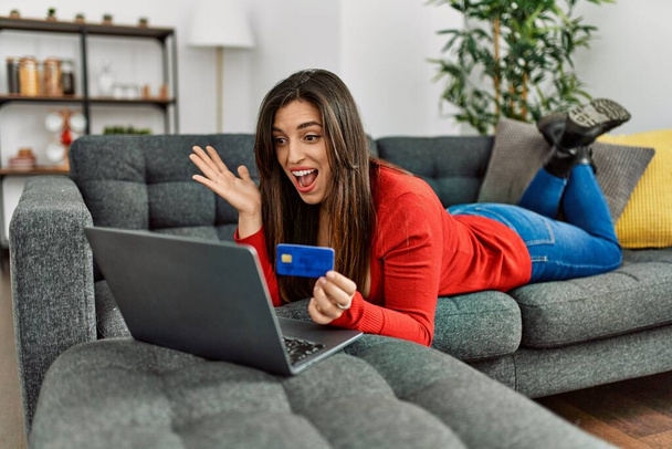 Giovane donna con computer portatile e carta di credito sdraiata sul divano a casa - Foto, immagini
