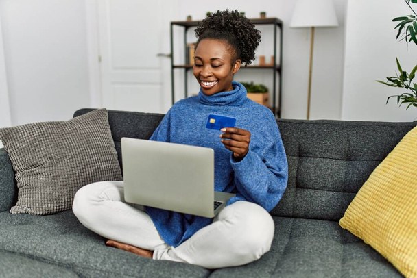 Afro-Amerikaanse vrouw met behulp van laptop en creditcard zittend op de bank thuis - Foto, afbeelding