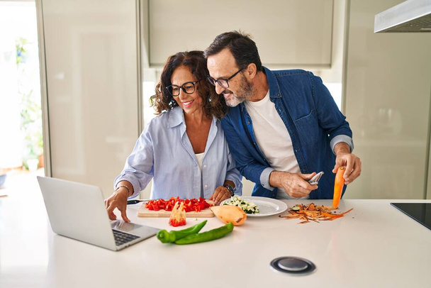 Orta yaşlı İspanyol çift mutfakta yemek pişiriyor ve laptop kullanıyor. - Fotoğraf, Görsel