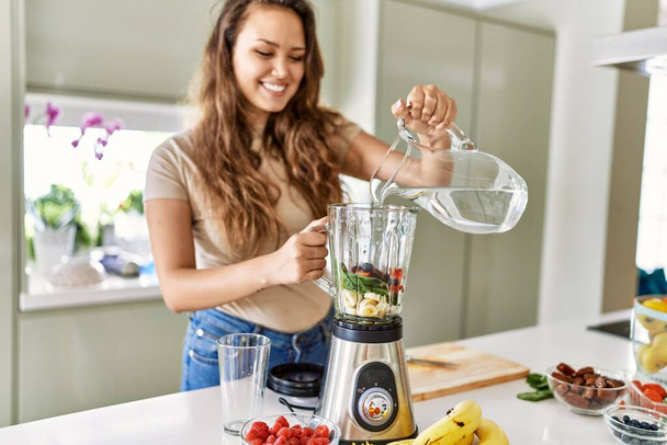 Young beautiful hispanic woman preparing vegetable smoothie pouring water on blender at the kitchen - 写真・画像