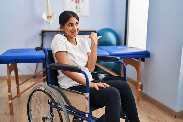 Young hispanic woman sitting on wheelchair at physiotherapy clinic pointing thumb up to the side smiling happy with open mouth  - Photo, Image