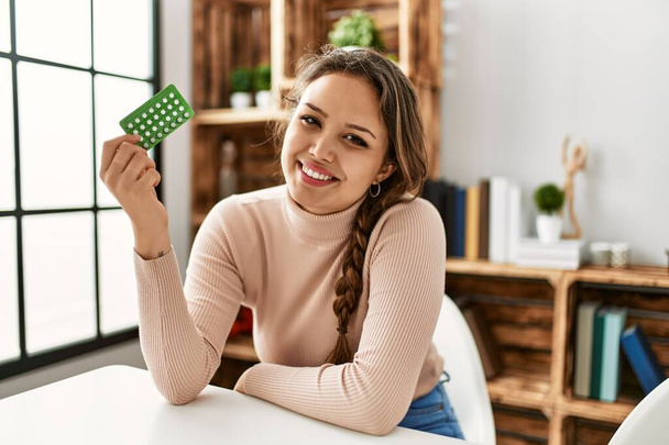 Young beautiful hispanic woman holding birth control pills sitting on table at home - Fotografie, Obrázek