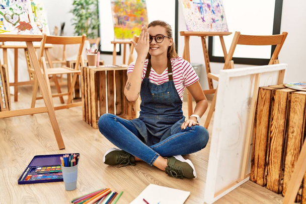 Νεαρή μελαχρινή γυναίκα στο στούντιο τέχνης κάθεται στο πάτωμα καλύπτοντας το ένα μάτι με το χέρι, με αυτοπεποίθηση χαμόγελο στο πρόσωπο και συγκίνηση έκπληξη.  - Φωτογραφία, εικόνα