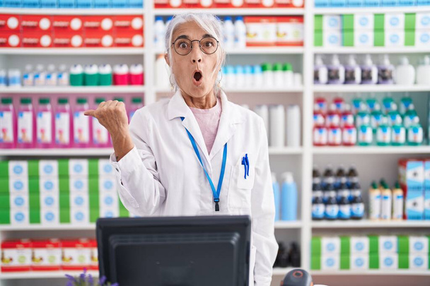 Mujer de mediana edad con tatuajes que trabajan en farmacia farmacia sorprendida señalando con el dedo de la mano a un lado, boca abierta expresión asombrada.  - Foto, Imagen