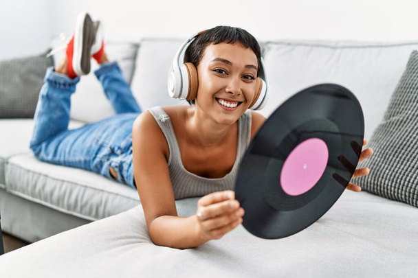 Young hispanic woman listening to music holding vinyl disc lying on sofa at home - 写真・画像