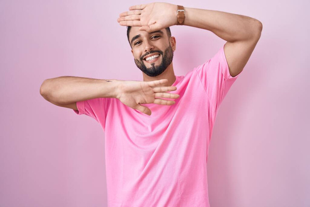 Latino nuori mies seisoo vaaleanpunainen tausta hymyillen iloinen pelaa kurkistaa boo kädet osoittavat kasvot. yllättynyt ja poistunut  - Valokuva, kuva