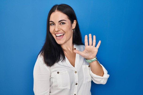Junge hispanische Frau steht vor blauem Hintergrund und zeigt mit Finger Nummer fünf nach oben, während sie selbstbewusst und glücklich lächelt.  - Foto, Bild