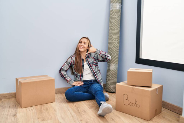 Young blonde woman sitting on the floor moving to a new home smiling doing phone gesture with hand and fingers like talking on the telephone. communicating concepts.  - Photo, Image