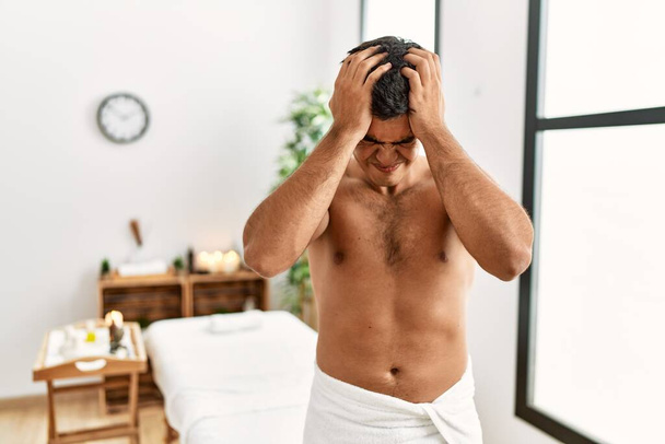 Young hispanic man standing shirtless at spa center suffering from headache desperate and stressed because pain and migraine. hands on head.  - Zdjęcie, obraz