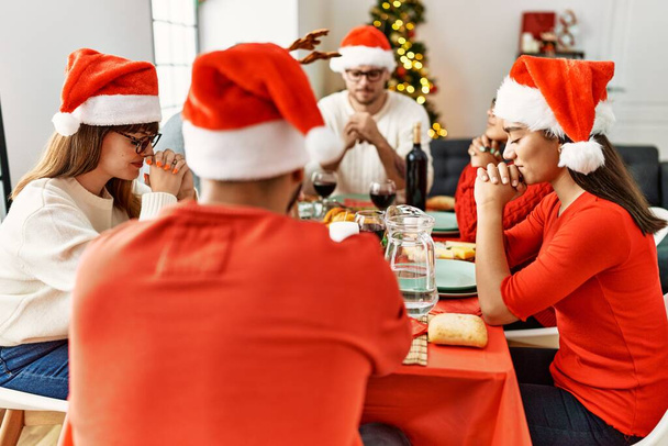 クリスマスを祝う若者のグループ自宅でテーブルの上に座って食べ物を祈る. - 写真・画像