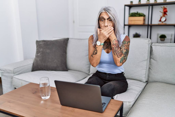 Grauhaarige Frau mittleren Alters, die zu Hause ihren Laptop benutzt, schockiert, als sie fälschlicherweise den Mund mit den Händen bedeckt. Geheimes Konzept.  - Foto, Bild